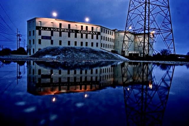 the J.J. Koepsell Company building at blue dusk, mirrored in the river, transmission tower alongside. Second image for #62, joins the tugboat.