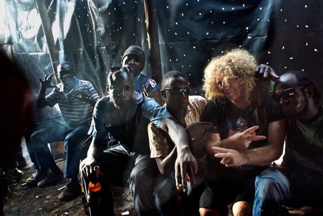 Mike with a group of men in a corrugated shelter, Africa travel, peace signs