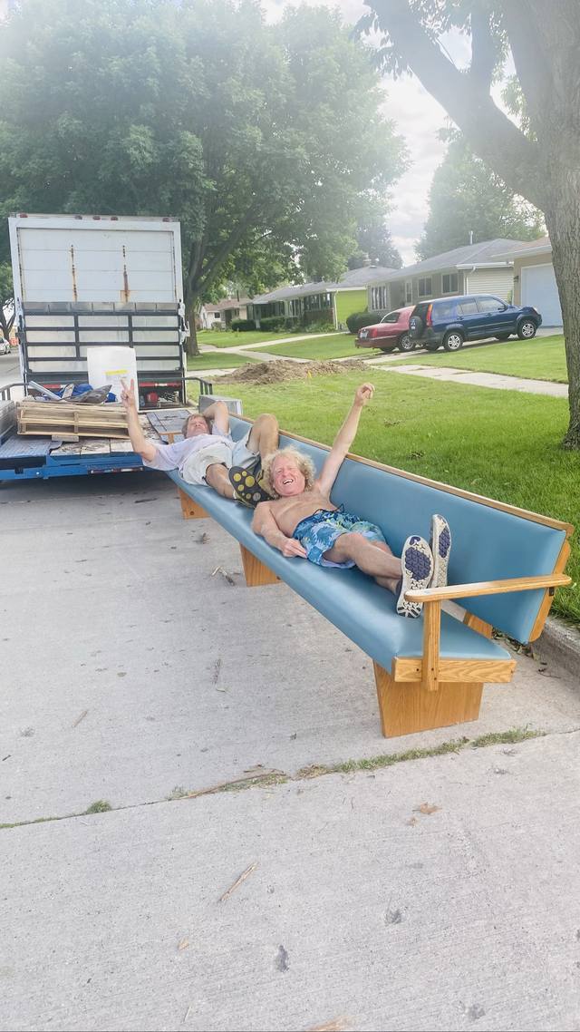 Mike and a friend sprawled on a long blue bench, moving truck behind, peace signs