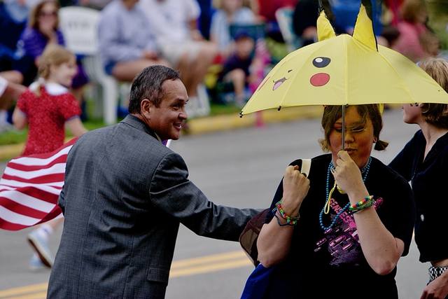 former Mayor **Juan Perez** (52nd Mayor of Sheboygan, 2005–2009; first Latino mayor in Wisconsin — verified via Wikipedia) in a suit with an American flag cape, beside a girl with a Pikachu umbrella. Mike ID'd him 2026-09-25.