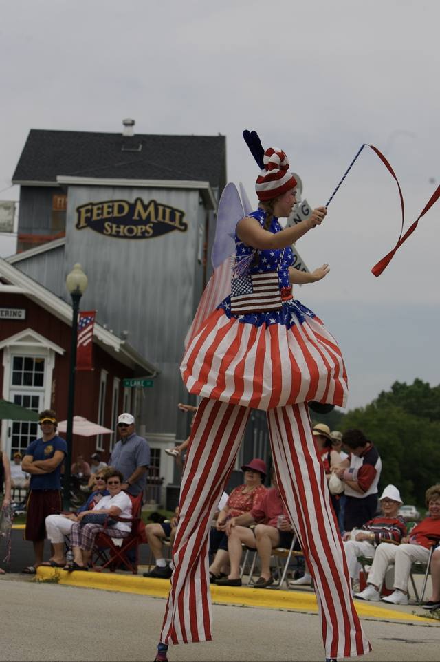 stilt-walking performer in a patriotic costume, Feed Mill Shops behind (parade set