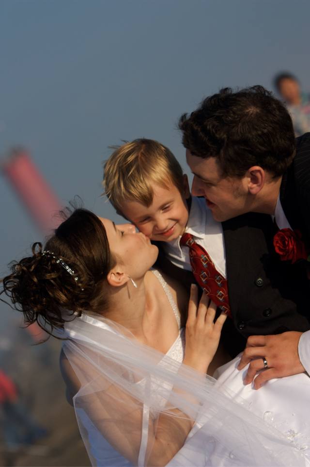 Bride kissing the little ring-bearer, groom leaning in — wedding on the breakwater